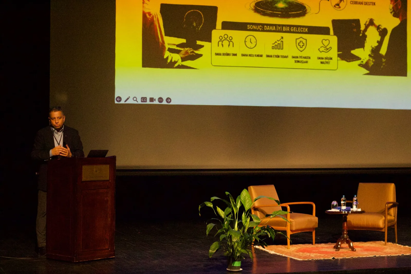 A speaker presenting from the lectern, with a slide on the screen and two armchairs on the stage
